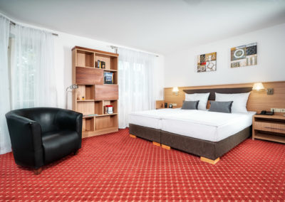 Doppelzimmer mit Doppelbett, Ledersessel und Bücherregal vor hellen Fenstern | Apart Business Hotel